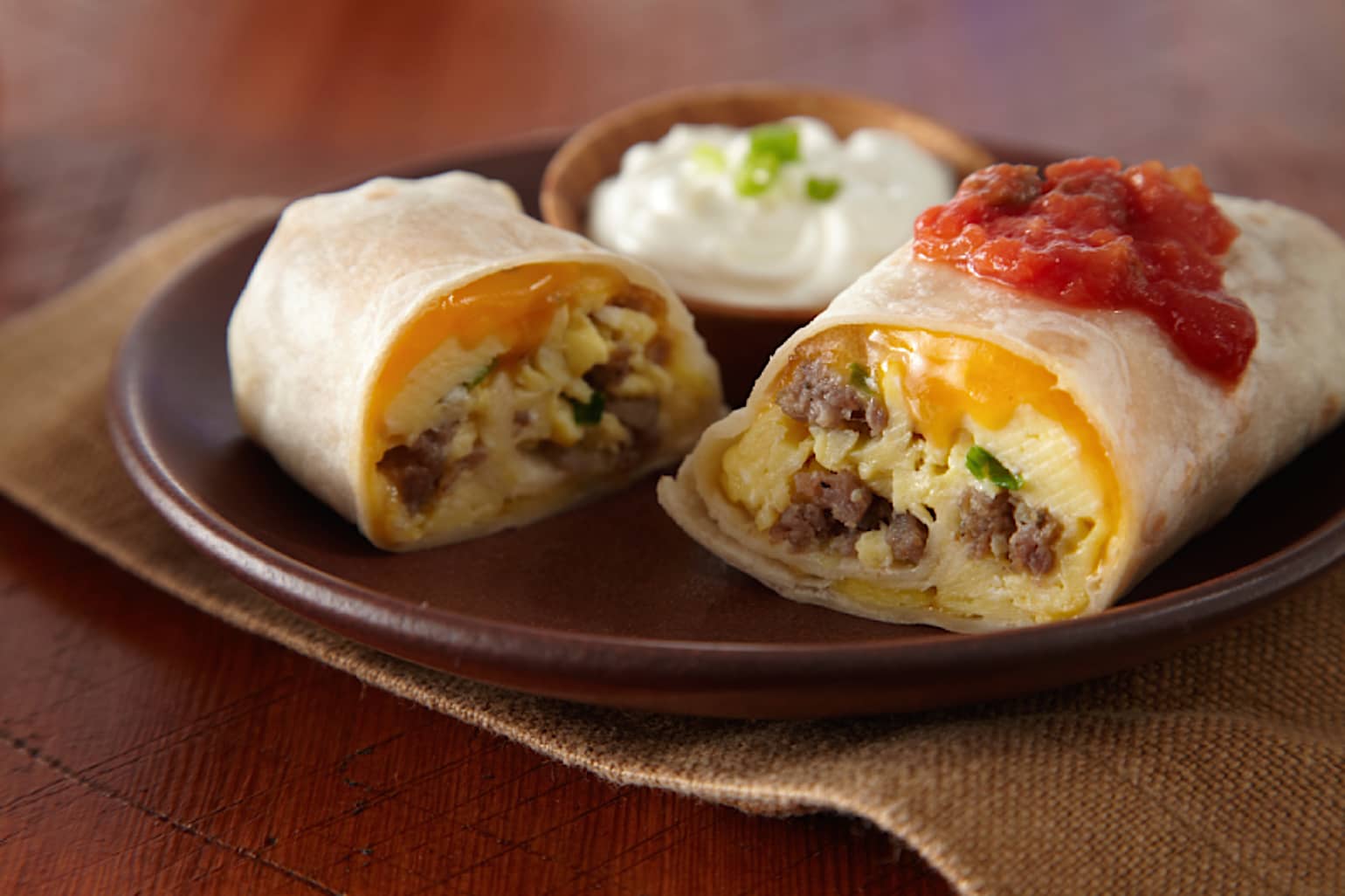 Un burrito de petit-déjeuner aux œufs, saucisses et fromage coupé en deux avec de la salsa et de la crème sure sur une assiette brune.